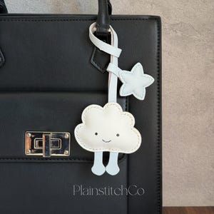 May include: A black handbag with a white leather cloud and star charm. The cloud has a smiling face and light blue legs. The charm is attached to the bag's handle with a white strap. The text "PlainstitchCo" is visible.