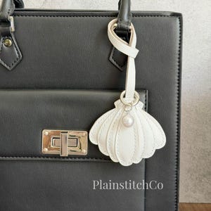 Puede incluir: Un llavero blanco en forma de concha con un acento de perla cuelga de un bolso negro. El llavero está sujeto al asa del bolso con una correa de cuero blanco y un anillo plateado. El bolso tiene un cierre plateado.