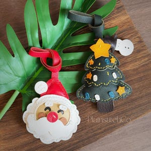 Puede incluir: Dos adornos navideños de cuero: un Papá Noel y un árbol de Navidad. El adorno de Papá Noel tiene un gorro rojo, barba blanca y una cara sonriente. El árbol de Navidad es verde oscuro con detalles de estrellas amarillas. Ambos tienen lazos de cuero para colgar.