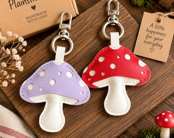 May include: Two mushroom-shaped keychains made of textured material, one in lavender and one in red, both featuring white spots and white stems. Each keychain has a silver-toned metal swivel clasp and ring attached to a white loop at the top.