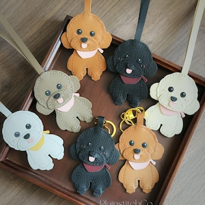 May include: A selection of poodle-shaped keyrings crafted from leather in shades of tan, black, and cream. Each keyring has a stitched design, a pink or red scarf, and a matching leather strap or gold keyring.
