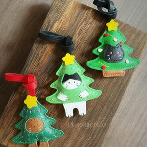 May include: Three green Christmas tree-shaped ornaments with cat faces. Each ornament has a yellow star on top and a leather loop. One cat is black, one is brown, and one is black and white. The ornaments are on a wooden surface.