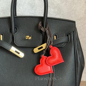 Puede incluir: Un bolso de mano de cuero negro con herrajes dorados y dos colgantes rojos en forma de corazón colgando del asa. El bolso tiene una superficie texturizada y un diseño clásico.
