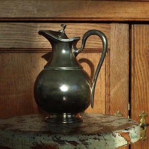 May include: A silver-colored pitcher with a rounded body, a curved handle, and a spout. The pitcher has a lid with a small decorative element on top. It sits on a small, round, distressed wooden table. The background is a wooden cabinet.