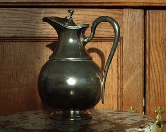 Op de afbeelding: Een zilverkleurige kan met een afgeronde body, een gebogen handvat en een tuit. De kan heeft een deksel met een klein decoratief element bovenop. Hij staat op een kleine, ronde, verweerde houten tafel. De achtergrond is een houten kast.