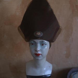 Może przedstawiać: Vintage brązowy kapelusz z wysoką, spiczastą koroną i ozdobną broszką. Kapelusz jest wystawiony na głowie manekina z czerwoną szminką i niebieskim cieniem do powiek. Manekin ma białą twarz i czarno-żółtą sukienkę.