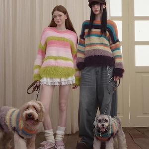 May include: Two people in colourful, striped sweaters, with dogs in matching sweaters. The sweaters have horizontal stripes in pink, green, orange, and purple. The dogs wear similar sweaters. The background includes a door and a neutral-coloured curtain.