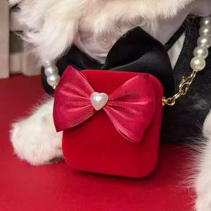 Pode incluir: Uma pequena caixa de veludo vermelho com um grande laço vermelho e uma pérola em forma de coração no centro. A caixa está presa a um colar de pérolas com um fecho dourado. O fundo inclui uma gravata borboleta preta e pele branca.