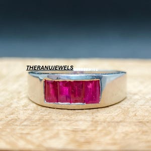 Peut inclure: Une bague en argent avec un sertissage rectangulaire. Le sertissage est orné de cinq pierres rouge rubis rectangulaires et vives. La bague est présentée sur une surface en bois. Le texte "THERANUJEWELS" est visible.