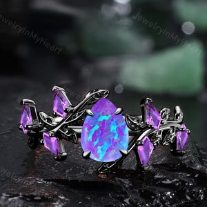 Puede incluir: Anillo negro con una piedra central en forma de pera, iridiscente azul-púrpura, rodeada de piedras moradas más pequeñas y un diseño de hojas. El anillo está sobre una superficie oscura y texturizada.