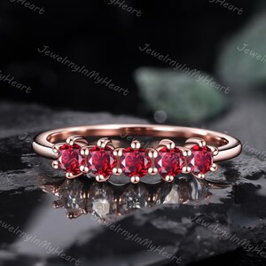 Könnte beinhalten: Ein roségoldener Ring mit fünf leuchtend roten Edelsteinen. Die Edelsteine sind in einer Reihe angeordnet. Das schmale Band des Rings betont die Edelsteine. Der Ring wird auf einer dunklen, strukturierten Oberfläche präsentiert. Der Ring ist etwa 0,5 cm breit.