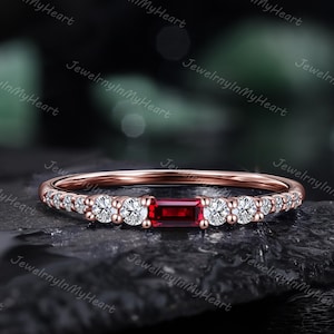 May include: A rose gold ring with a rectangular, deep red gemstone at the center, flanked by smaller, round, clear gemstones. The band is thin and adorned with additional clear gemstones. The ring is displayed on a dark, textured surface.