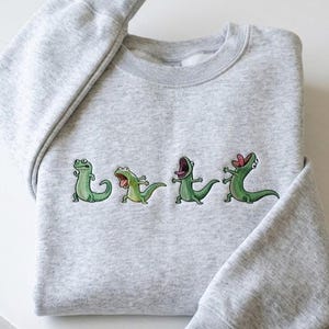 Puede incluir: Una sudadera gris claro con un diseño bordado de cuatro lagartos de dibujos animados verdes en varias poses. Los lagartos tienen la boca abierta y están en diferentes posiciones. La sudadera está doblada cuidadosamente sobre una superficie blanca.