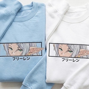 Può includere: Due felpe, una azzurra e una bianca, ciascuna con una grafica ricamata in stile anime. La grafica è un design rettangolare con gli occhi di un personaggio e il testo giapponese "フリーレン".