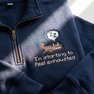 May include: A navy blue quarter-zip sweatshirt featuring an embroidered pixel art design of a brown cat sleeping with a white speech bubble containing the letters ZZZ. Below the cat, the text reads, I'm starting to feel exhausted.