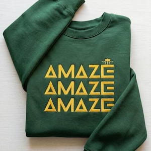 Könnte beinhalten: Ein dunkelgrünes Sweatshirt mit dem Wort "AMAZE" in gelben Blockbuchstaben, dreimal gestapelt. Eine kleine gelbe Grafik befindet sich über dem oberen Wort. Das Sweatshirt ist auf einer weißen Oberfläche gefaltet.