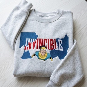 Może przedstawiać: Jasnoszara bluza z napisem "INVINCIBLE" w czerwono-białych literach, na tle niebieskiego konturu stanu. Poniżej znajduje się rysunkowy obrazek superbohatera w niebiesko-żółtym kostiumie.
