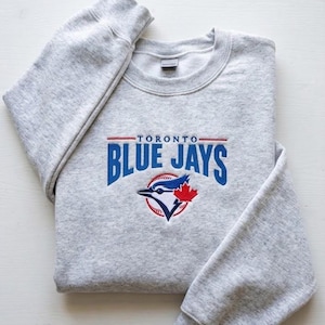 Può includere: Una felpa grigio chiaro con la scritta "TORONTO BLUE JAYS" in blu e rosso. La felpa presenta il logo dei Blue Jays, un uccello ghiandaia azzurra con una foglia d'acero, sul davanti.