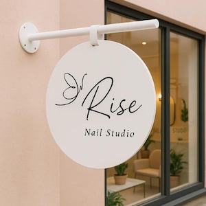 Op de afbeelding: Een wit, rond bord voor "Rise Nail Studio" hangt aan een witte metalen beugel. Het bord heeft een elegant zwart schrift en een vlinderontwerp. Het bord is bevestigd aan een lichtroze buitenmuur, met een raam op de achtergrond.