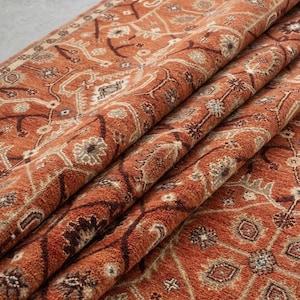 May include: A folded, terracotta-coloured rug with a detailed, traditional pattern. The design incorporates floral and geometric elements in shades of brown, cream, and blue. The rug's texture appears soft and plush, with visible folds adding depth and dimension.