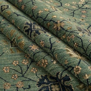 May include: Close-up of a rolled-up, teal-colored rug with a detailed floral pattern. The rug features a dark blue and tan floral design on a textured surface. The rug's design includes small flowers and vines.