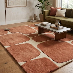 May include: A terracotta-coloured area rug featuring an abstract design of large, rounded shapes set against a cream backdrop. The rug is in a living room with a green sofa, coffee table, and potted plants. The rug's design adds a modern touch to the room.