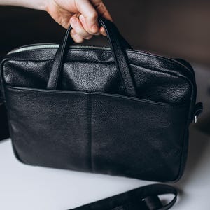 Op de afbeelding: Een zwarte leren aktetas met een getextureerd oppervlak, met een voorvak en een zilveren ritssluiting. De tas heeft twee handvatten en een afneembare schouderriem. De aktetas wordt door een hand vastgehouden.
