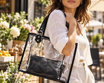 May include: A clear rectangular tote bag with black trim and black shoulder straps. Inside the transparent bag is a solid black rectangular pouch. The bag features silver-toned metal hardware connecting the straps to the main body.