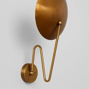 May include: A gold-colored wall sconce with a large, round, semi-circular shade. The shade is connected to a curved arm and a circular base, all in the same metallic finish. The design is minimalist and modern.