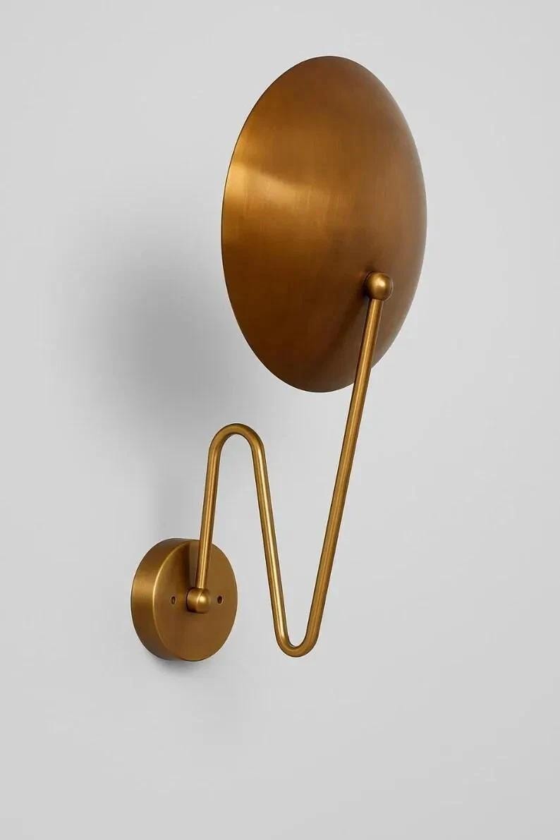 May include: A gold-colored wall sconce with a large, round, semi-circular shade. The shade is connected to a curved arm and a circular base, all in the same metallic finish. The design is minimalist and modern.