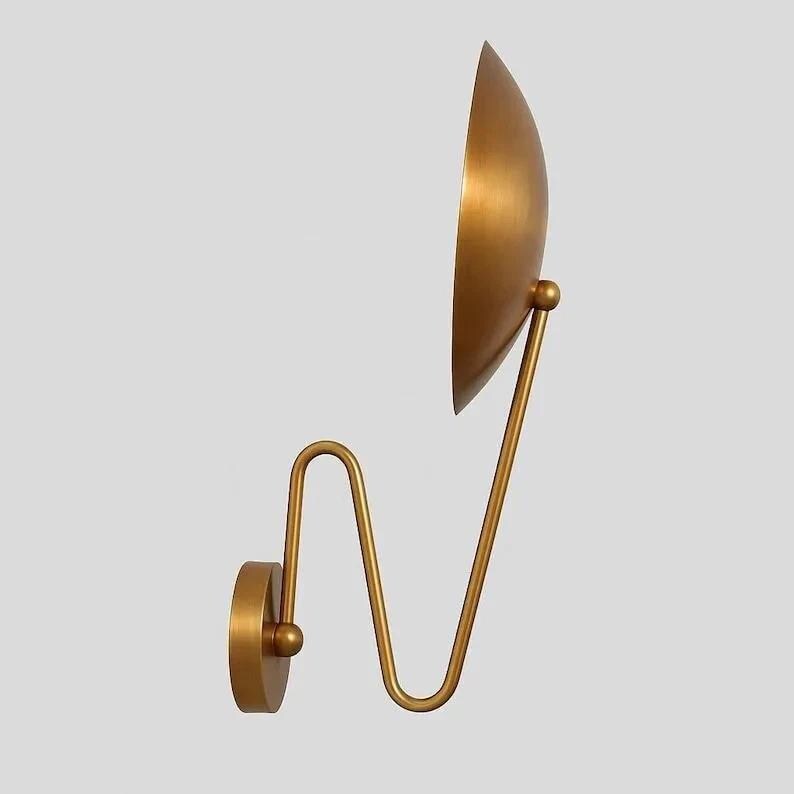 May include: A gold-colored wall sconce with a semi-circular shade and a curved arm. The shade is positioned to the side, and the arm has a unique, decorative shape. The sconce is mounted on a circular base.