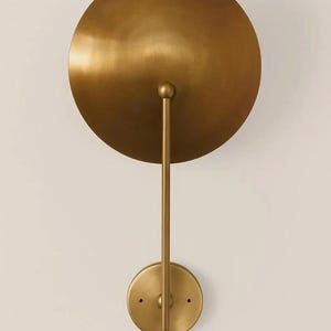May include: A gold-colored wall sconce with a large, circular shade and a smaller, circular base. The shade is connected to the base by a straight, vertical rod. The sconce has a minimalist, modern design.
