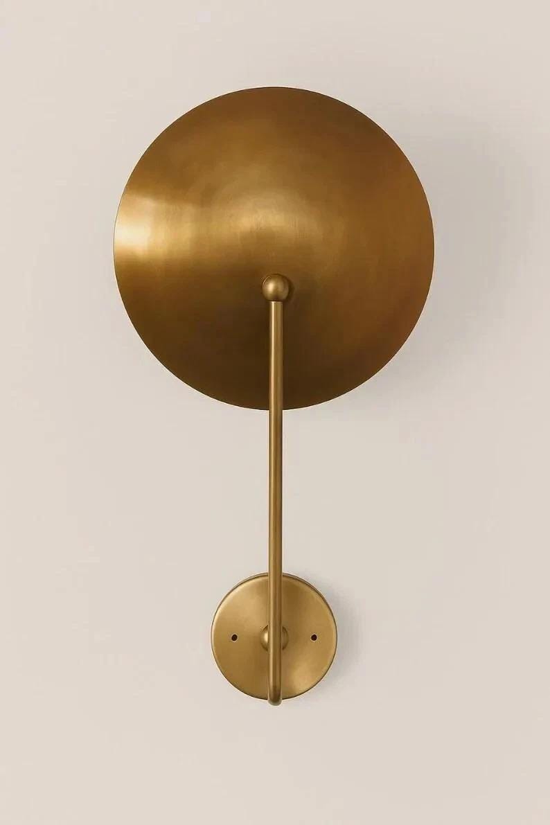 May include: A gold-colored wall sconce with a large, circular shade and a smaller, circular base. The shade is connected to the base by a straight, vertical rod. The sconce has a minimalist, modern design.