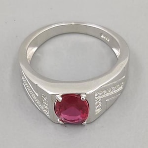 Op de afbeelding: Een zilveren ring met een rechthoekige voorkant en een grote, ronde, rode edelsteen. De ring heeft een eenvoudig, elegant ontwerp met kleine decoratieve details aan de zijkanten. De edelsteen is het middelpunt van de ring.