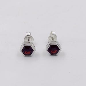 May include: A pair of silver stud earrings. Each earring features a hexagonal, dark red gemstone set in a silver bezel. The earrings are displayed against a plain white background.