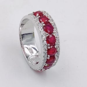 Könnte beinhalten: Ein silberner Ring mit einem Band aus abwechselnd roten Edelsteinen und kleineren, klaren Edelsteinen. Der Ring ist vor einem weißen Hintergrund platziert. Der Ring hat die Ringgröße 54 (EU).