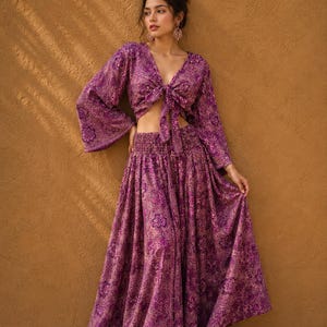 May include: A two-piece outfit consisting of a long-sleeved, V-neck crop top with a front tie closure and a matching high-waisted, floor-length maxi skirt with a smocked elastic waistband, both featuring a purple and magenta floral paisley print.