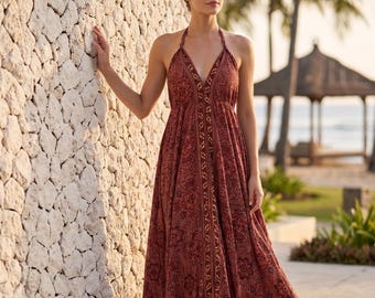 May include: A long, halter-neck dress in a reddish-brown hue with a paisley pattern. The dress features a V-neckline and a flowing skirt. The model is standing next to a stone wall, with a beach scene in the background.