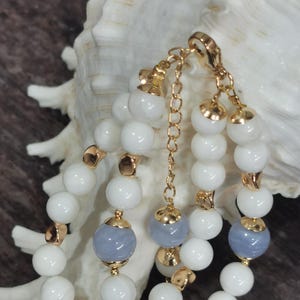Pu&ograve; includere: Bracciale a pi&ugrave; fili con perline bianche e grigio-blu, rifinito con dettagli in metallo color oro. Il bracciale &egrave; esposto su uno sfondo di conchiglia bianca e superficie marrone. Le perline sono rotonde e ben distanziate.