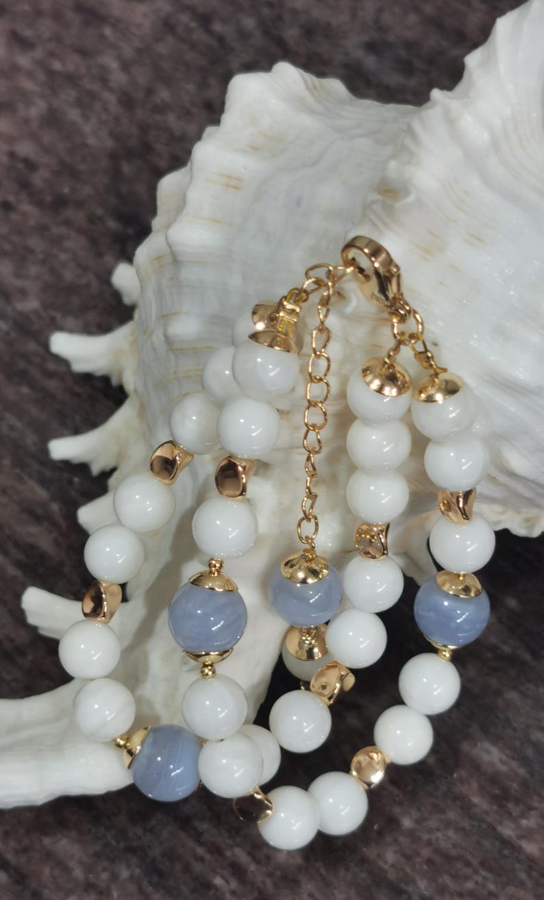 Pu&ograve; includere: Bracciale a pi&ugrave; fili con perline bianche e grigio-blu, rifinito con dettagli in metallo color oro. Il bracciale &egrave; esposto su uno sfondo di conchiglia bianca e superficie marrone. Le perline sono rotonde e ben distanziate.