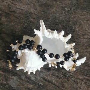 May include: A black beaded necklace with gold-coloured accents rests inside a white seashell. The necklace features matte black beads strung together with gold-toned metal. The seashell is white and has a textured surface.