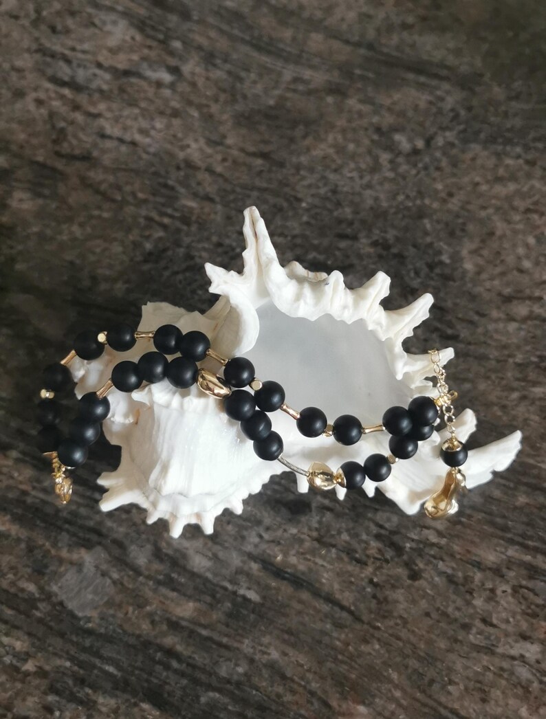 May include: A black beaded necklace with gold-coloured accents rests inside a white seashell. The necklace features matte black beads strung together with gold-toned metal. The seashell is white and has a textured surface.