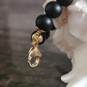 May include: A close-up of a black beaded bracelet with gold-coloured accents. The bracelet features matte black beads and a gold-coloured clasp. The jewellery is displayed against a blurred background.