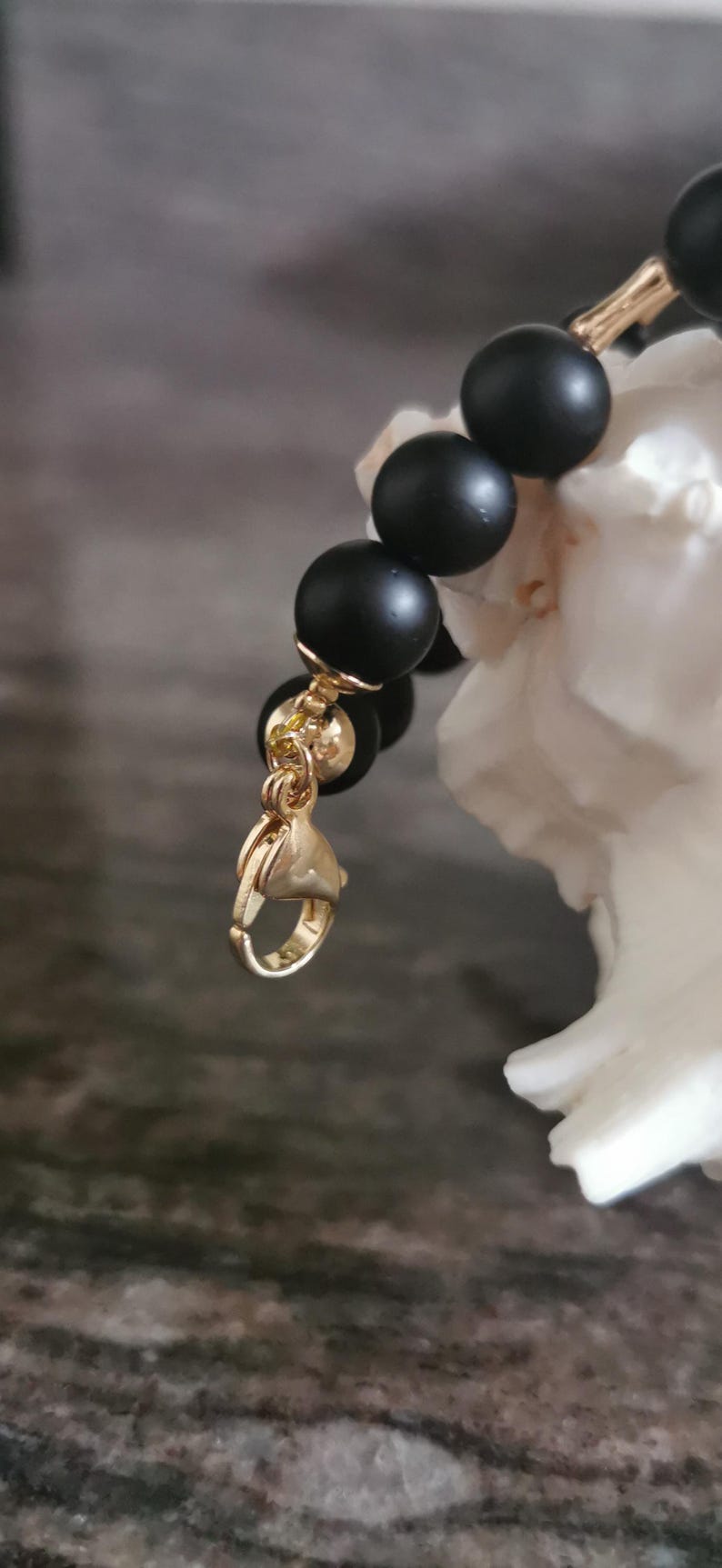 May include: A close-up of a black beaded bracelet with gold-coloured accents. The bracelet features matte black beads and a gold-coloured clasp. The jewellery is displayed against a blurred background.