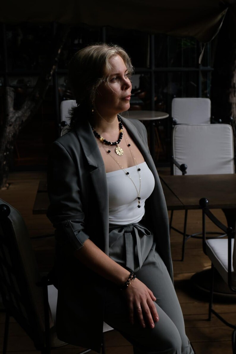 May include: A person wearing a grey blazer, white top, and grey trousers, accessorised with layered necklaces and a bracelet. The necklaces feature black beads and a gold pendant. The setting appears to be a restaurant or lounge with wooden tables and white chairs.