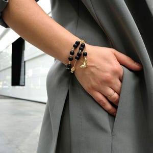 May include: A black beaded bracelet with gold detailing and a high-heeled shoe charm. The bracelet is worn on a wrist, with a grey blazer in the background. The bracelet is a fashion accessory.