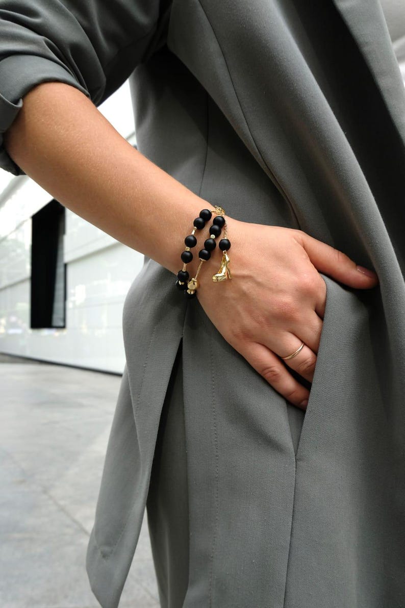 May include: A black beaded bracelet with gold detailing and a high-heeled shoe charm. The bracelet is worn on a wrist, with a grey blazer in the background. The bracelet is a fashion accessory.