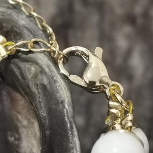 Pu&ograve; includere: Primo piano di una chiusura e catena di collana color oro, con una fila di grandi perle rotonde bianche. La chiusura collega la catena e le perle, creando un contrasto di colori e trame. Lo sfondo &egrave; un grigio scuro sfocato.