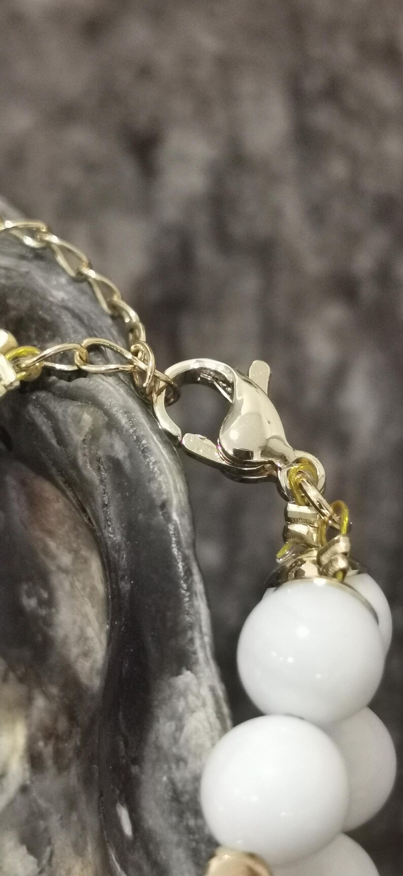 Pu&ograve; includere: Primo piano di una chiusura e catena di collana color oro, con una fila di grandi perle rotonde bianche. La chiusura collega la catena e le perle, creando un contrasto di colori e trame. Lo sfondo &egrave; un grigio scuro sfocato.