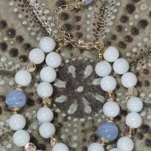 Pu&ograve; includere: Un set di collana e bracciale con perline bianche e azzurre. La collana ha una sottile catena d'argento, e entrambi i pezzi hanno dettagli color oro. I gioielli sono esposti su una superficie strutturata dai toni neutri.